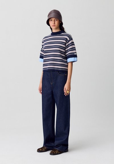 Suéter de manga corta a rayas en azul marino y celeste, combinado con jeans de denim oscuro de pierna ancha. Los accesorios incluyen un sombrero tipo bucket marrón y zapatos con estampado.