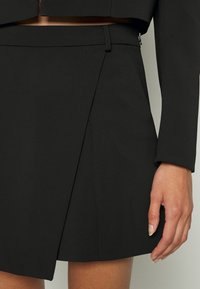 Närbild av en svart skräddarsydd kjol med ett framtill överlappande detalj, bärs med en matchande svart kort jacka på en modèles nedre torso.