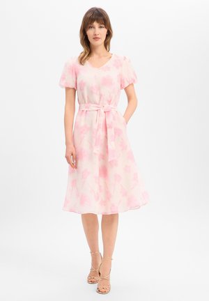 Femme portant une robe fleurie rose et blanche longueur genoux avec manches bouffantes et ceinture nouée à la taille, associée à des talons beiges à brides.