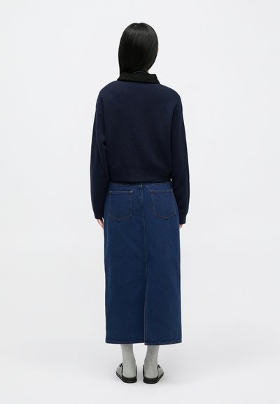 Μπλε πουλόβερ με ψηλό γιακά σε συνδυασμό με μακριά μπλε τζιν φούστα, που διαθέτει δύο πίσω τσέπες και ίσια γραμμή. Το σύνολο ολοκληρώνουν γκρι κάλτσες και μαύρα σανδάλια.