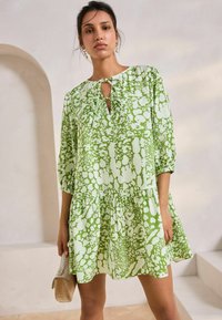 Rochie verde cu un model, croi lejer, mâneci trei sferturi și decolteu în V cu un detaliu legat. Țesătură texturată cu fustă din volane.