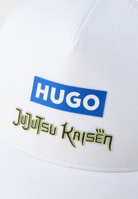 Gorra blanca con un diseño estructurado. Presenta un logo azul "HUGO" y texto en amarillo-verde "Jujutsu Kaisen" en la parte frontal. Textura de tela suave.