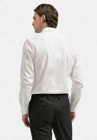 Vit skjorta med lång ärm, slätt bomullstyg, strukturerad krage och franska manschetter. Bärs med mörka byxor, bakifrån.