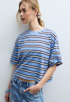 Jeune femme aux cheveux blonds attachés en arrière avec un chouchou, portant un t-shirt à rayures bleu et un jean en denim bleu clair, debout devant un mur blanc.