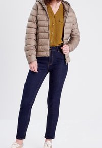 Femme portant une doudoune beige sur une chemise à boutons moutarde, jean skinny bleu foncé et baskets blanches, se tenant devant un fond uni.