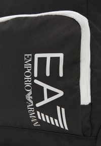 Borsa nera in tessuto con dettagli bianchi e un logo che recita "EA Emporio Armani" con linee grafiche, enfatizzando un design moderno.
