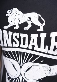 Zwarte T-shirt met een witte leeuwgrafiek en bokshandschoenen, met vetgedrukte tekst "Lonsdale." Het ontwerp heeft een versleten, textuurlijk uiterlijk.