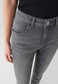 Primo piano di una persona che indossa jeans in denim grigi sbiaditi con una maglietta nera, che mostra le tasche anteriori, i passanti per la cintura e la chiusura a bottone.