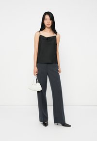 Frau in einem schwarzen Satin-Cami-Top, dunklen gestreiften weiten Hosen, schwarzen spitzen High Heels, die eine kleine weiße Raffungstasche hält.