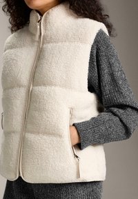 Frau trägt eine cremefarbene Sherpa-Fleece-Weste über einem dunkelgrauen, langärmligen Stricktop, die Hand in der Reißverschlusstasche der Weste.