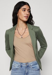 Blazer verde ajustat, dintr-un material moale, purtat peste un top bej. Modelul poartă blugi bleu deschis și un colier gros din argint.
