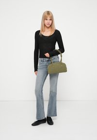 Sac à main en tissu vert de forme rectangulaire, avec deux courtes poignées et de petits détails en métal argenté. Le modèle porte un haut noir et un jean évasé.