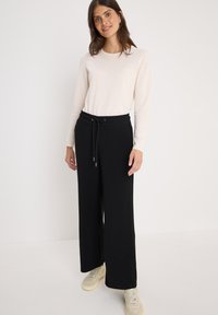 Pantalons noirs à jambes larges avec une taille élastique et un cordon de serrage, associés à un haut beige clair à manches longues, portés avec des baskets de couleur claire.