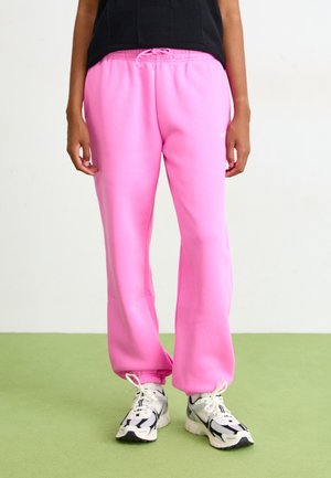 Person wearing bright pink sweatpants with white and black sneakers standing on green floor against white background.