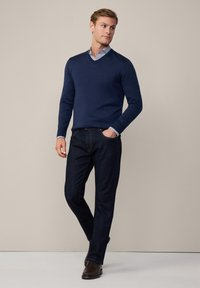 Maglione navy a collo a V con maniche lunghe, abbinato a jeans in denim scuri e scarpe marroni; sovrapposto a una camicia leggera con bottoni sotto.