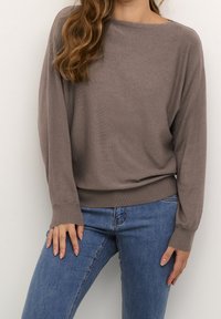 Maglione marrone a maniche lunghe con scollatura ampia a barca, polsini e orlo a coste. Abbinato a jeans skinny blu e mostrato su uno sfondo neutro.