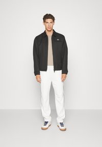 Chaqueta negra con cuello, camisa beige, pantalones blancos con rayas laterales y zapatillas blancas con suelas marrones, de pie sobre un fondo simple.