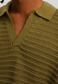Gros plan sur une personne portant un pull en maille vert olive avec un col et un décolleté en V, montrant les détails de la texture du tissu.