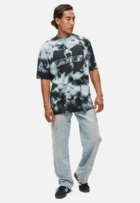 Černé a šedé tričko s tímovým vzorem tie-dye s velkým logem Wu-Tang, v kombinaci se světle modrými roztrhanými džíny a černými botami.