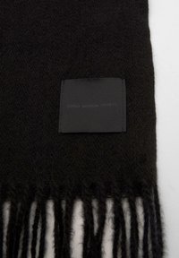 Črni volneni šal z resicami. Ima kvadratno oznako, prišito v tkanino, na kateri stoji napis "KLASIK MODERNO VAŠE".