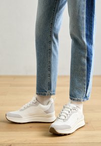 Zapatillas deportivas blancas con acentos grises y parte superior texturizada, combinadas con jeans azul claro remangados, sobre un fondo de suelo de madera.