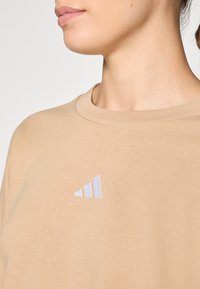 Beige sweatshirt med rund halsringning og et hvidt broderet tre-stribe logo design på venstre side. Glat tekstur.