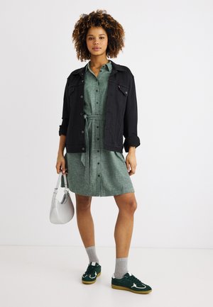 Woman wearing a green buttoned dress with a waist tie, black jacket, gray socks, green sneakers, and holding a white handbag.