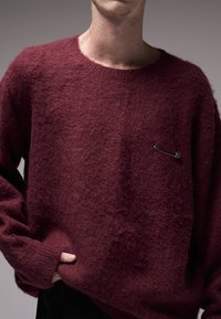 Μπλουζάκι σε μπορντό χρώμα, φτιαγμένο από αφράτο υλικό, με στρογγυλή λαιμόκοψη και διακοσμητικό τσακιστό καρφί ασφαλείας στο μπροστινό μέρος.