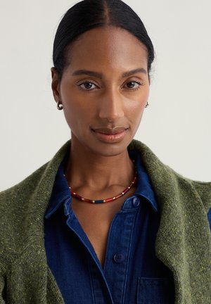 Collana di perline colorata con accenti rossi, blu e dorati, indossata con un cardigan verde a trama su una camicia di jeans blu.