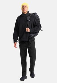 Jonge man met een gele beanie, zwart fleecejack, zwarte broek en wandelschoenen draagt een grijze rugzak over één schouder.