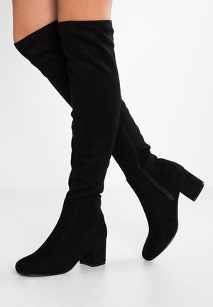 Bottes en daim noir au-dessus du genou avec talons blocs et fermetures éclair latérales, portées sur des jambes croisées sur un fond blanc uni.