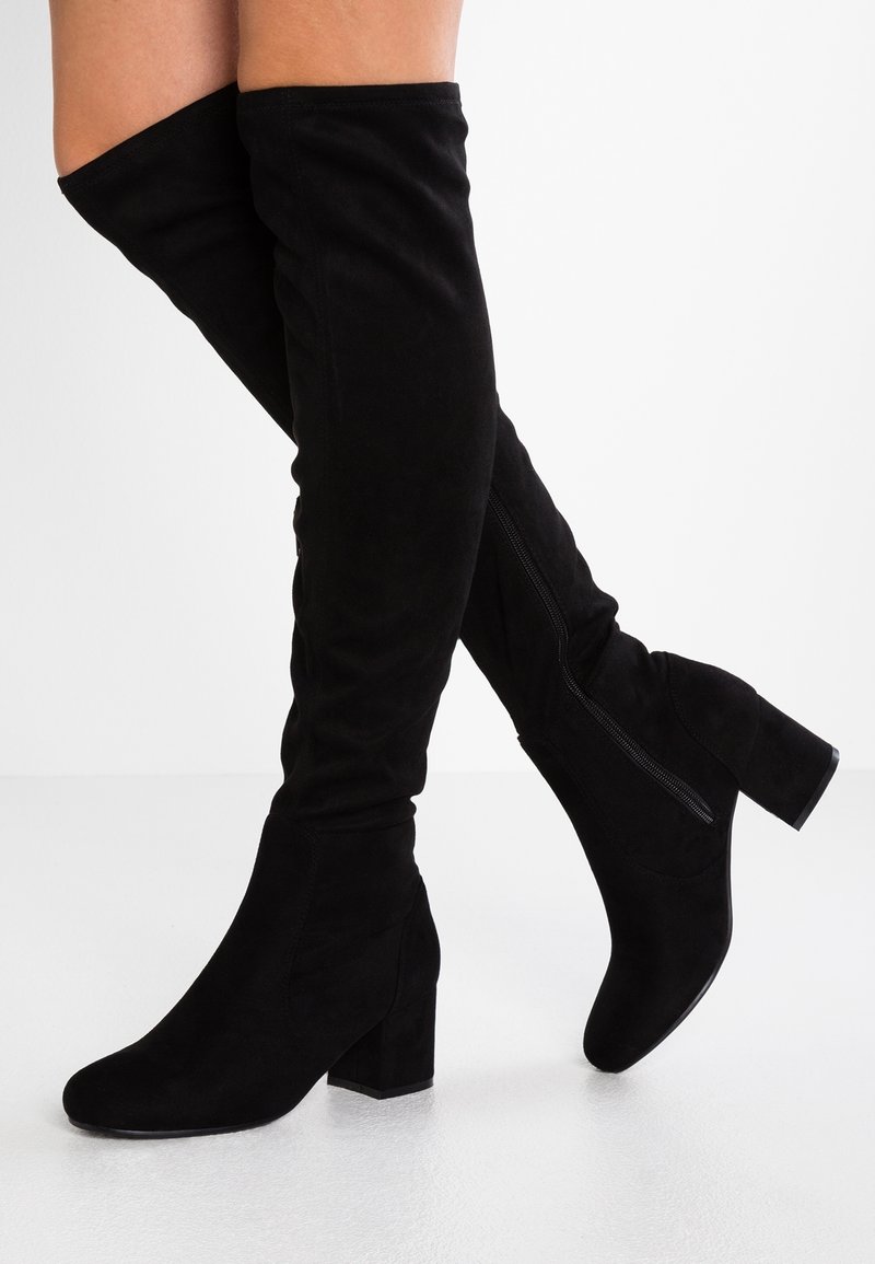Bottes en daim noir au-dessus du genou avec talons blocs et fermetures éclair latérales, portées sur des jambes croisées sur un fond blanc uni.