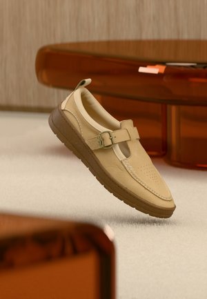 Beige leather slip-on shoe with buckle strap and brown rubber sole, positioned mid-air above light carpeted floor and near amber translucent furniture.