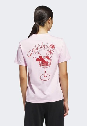 Camiseta de algodón rosa claro con un gráfico rojo en la parte posterior, que muestra a una mujer recostada en una copa de margarita con el texto "Adidas".