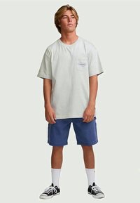 Jovem em pé, vestindo uma t-shirt cinza-claro com bolso, calções azuis, meias brancas e ténis pretos, virado ligeiramente para a direita.