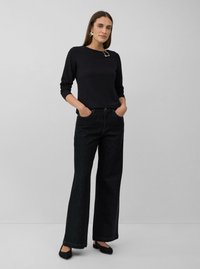 Vrouw draagt een zwart lange mouwen top met een decoratieve gesp, zwarte wijde jeans, zwarte platte schoenen en vlinder-vormige oorbellen.