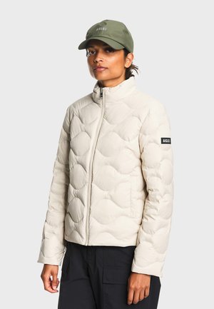 Femme portant une veste matelassée beige clair avec un col montant et une casquette verte Aigle, debout devant un fond clair uni.
