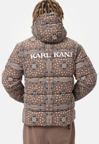 Brązowa kurtka puchowa z wzorem paisley i kwiatowym, z kapturem i białym napisem "KARL KANI" na plecach.