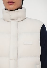 Helle beige gesteppte Weste mit hohem Kragen, ausgestattet mit einem Druckknopfverschluss vorne und einem kleinen Logopatch auf der linken Seite. Weicher, glatter Stoff.