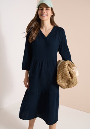 Uśmiechnięta kobieta w granatowej sukience midi i jasnoniebieskiej czapce trzymająca dużą plecioną słomianą torbę na beżowym i białym tle.