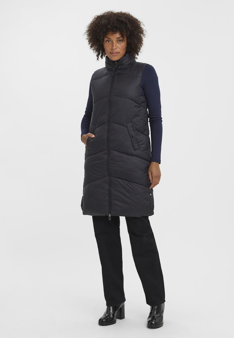 Juodas paminkštintas liemenė su aukštu kaklu ir priekiniais užtrauktukais, su šoninėmis kišenėmis ir siuvinėtu dizainu, dėvima virš tamsiai mėlynos ilgomis rankovėmis marškinėlių ir juodų kelnių.