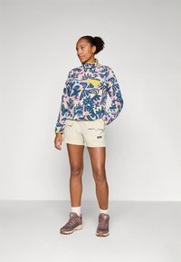 Fleecinis megztinis su spalvingu gėlių raštu, pusiau užtrauktuku ir geltonu kišeniu. Kreminės spalvos šortai, rudi žygių batai ir balti kojinės papildo aprangą.