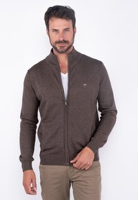 Brun zip-up genser laget av strikket stoff, med ståkrage og ribbestrikkede mansjetter, båret over en hvit T-skjorte. Subtil logo detalj på brystet.