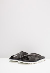 Sandali neri in pelle con cinturini incrociati e suola testurizzata. Presentano una suola bianca seghettata e linee di accento grigio sottili.