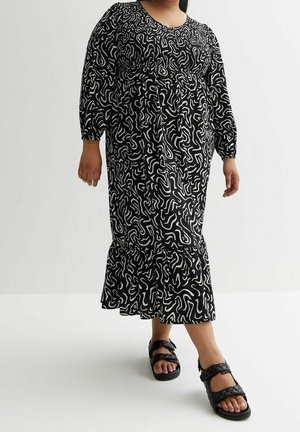 Kvinde iført en lang kjole med sort og hvidt abstrakt mønster og sorte quiltede sandaler, stående mod en ensartet lys baggrund.