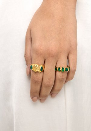 Ręka z dwoma złotymi pierścionkami o nieregularnych kształtach i zielonymi kamieniami inkrustowanymi, noszonymi na palcu środkowym i serdecznym na białym tle.