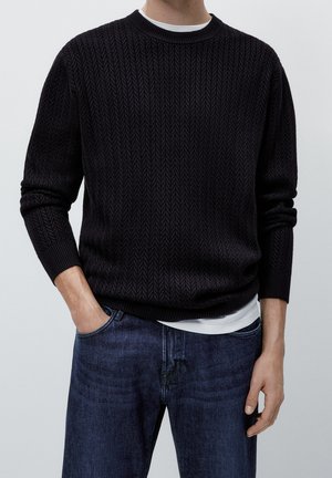 Mężczyzna w czarnym, fakturowanym swetrze z okrągłym dekoltem, założonym na białą koszulę, wpuszczoną w ciemnoniebieskie dżinsy, z jedną ręką w kieszeni.