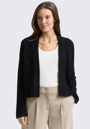 Black, textured cardigan with open front and long sleeves, worn over a white top. Paired with beige, pleated trousers.