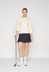 Ανοιχτό κρεμ πουλόβερ με ροζ γιακά, σε συνδυασμό με μαύρη φούστα γκολφ με πτυχώσεις. Φοράει λευκά αθλητικά παπούτσια και κρατάει ένα μπαστούνι γκολφ.