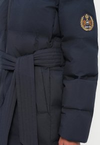 Marineblå pufferjakke med bælte i taljen, der har et struktureret stof, markante sømme og et broderet emblem på overarmen.